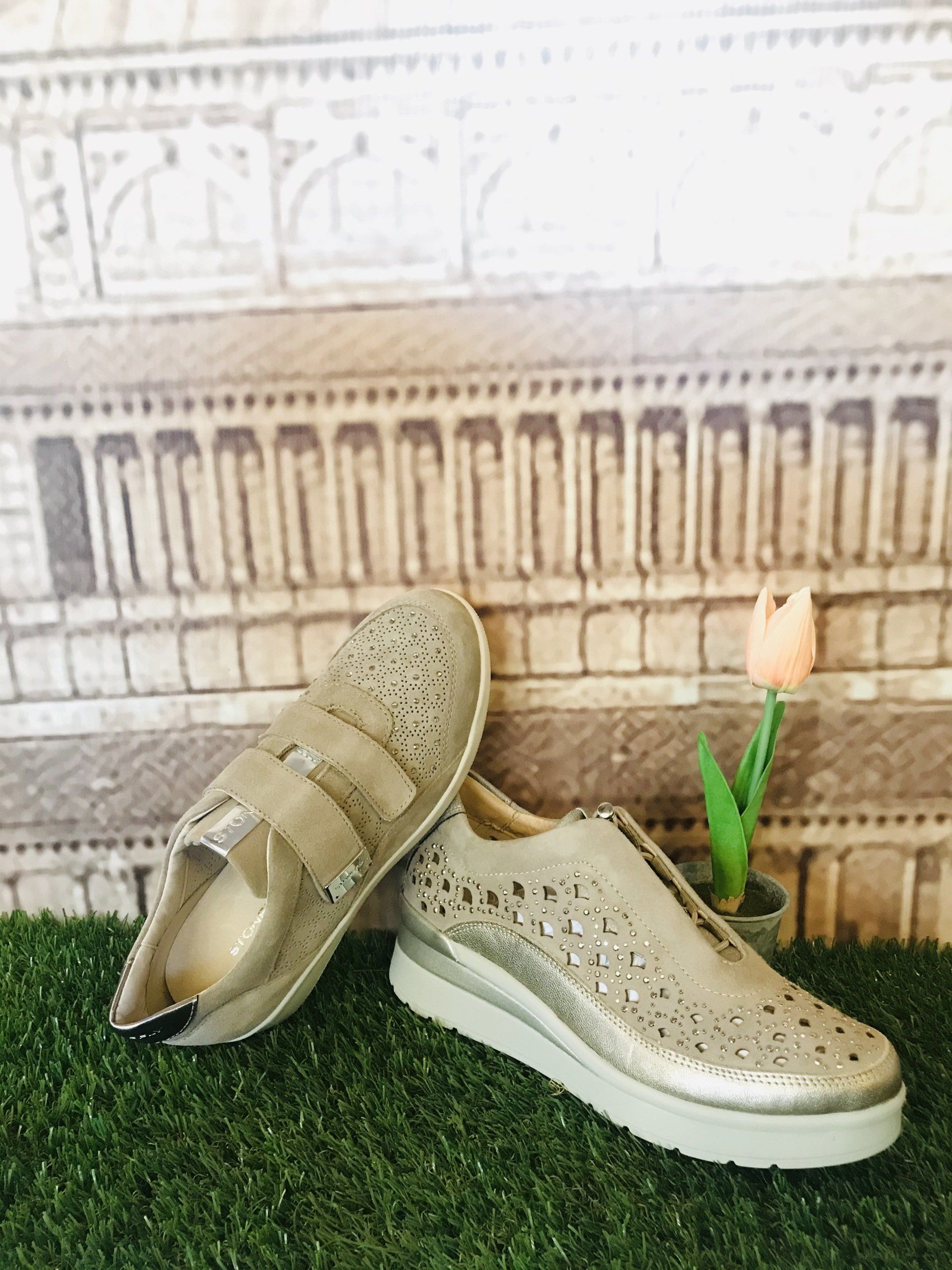 Sneakers argentate con dettagli intagliati e cinturini a strappo su erba sintetica verde, con uno sfondo di edifici e tulipani.