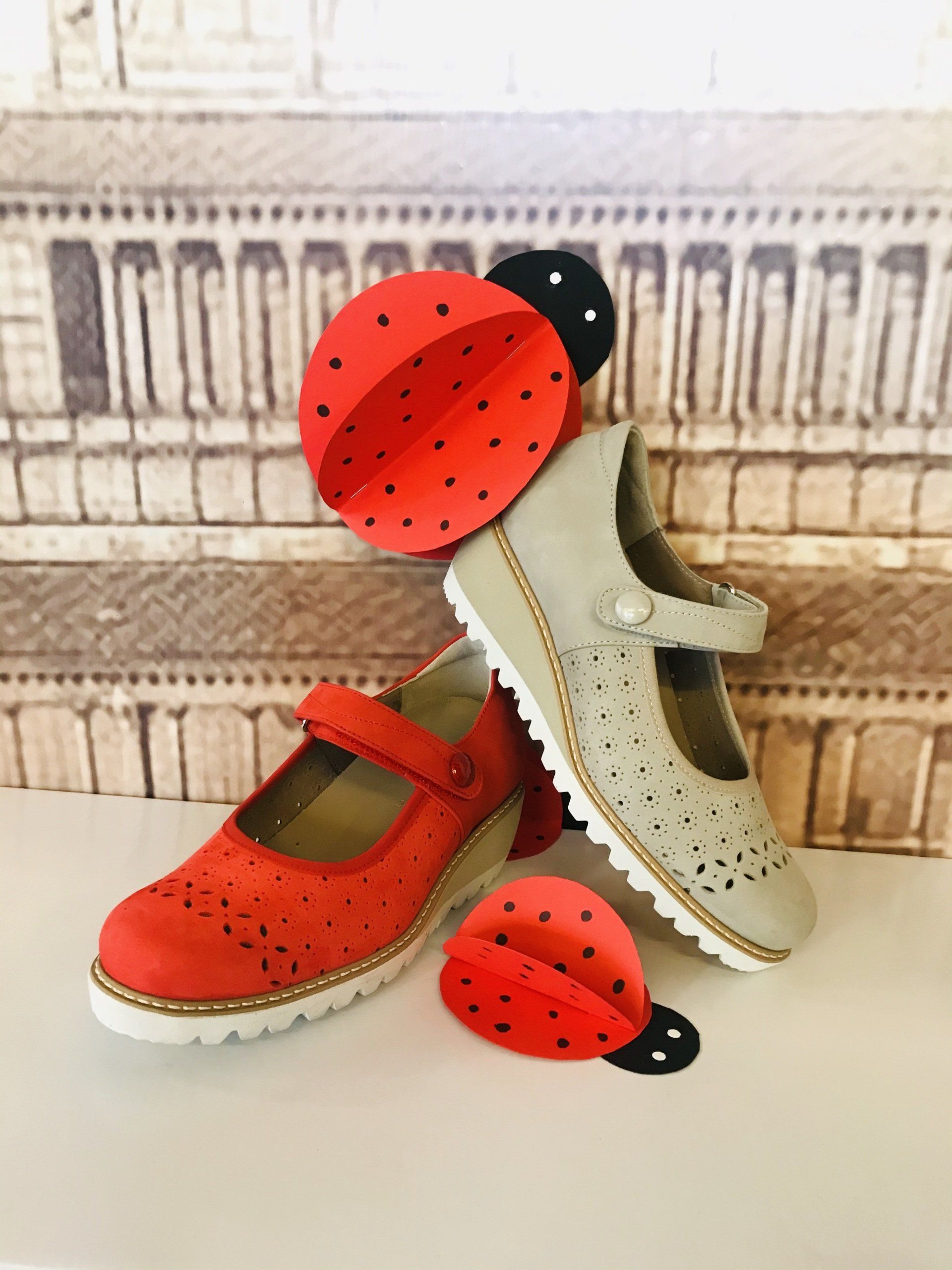 Scarpe per bambini rosse e beige con dettagli traforati, esposte con decorazioni a forma di coccinella sullo sfondo.