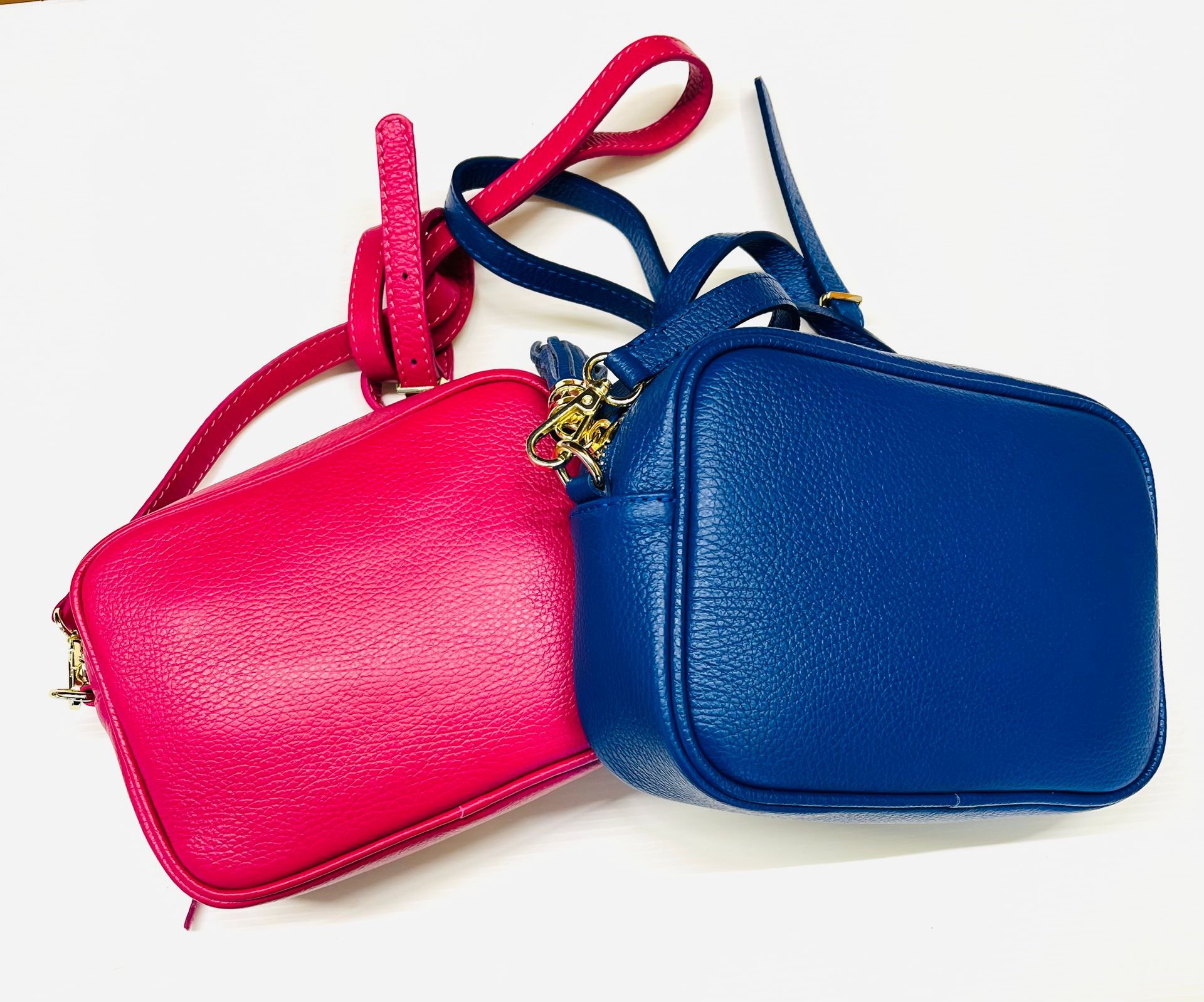 Due piccole borse a tracolla quadrate in pelle: una rosa e una blu, con lunghe tracolle e dettagli in catena dorata, su una superficie bianca.