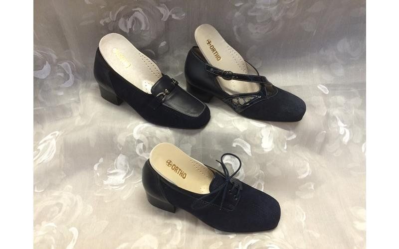 Tre paia di scarpe da donna in pelle scamosciata blu navy: un mocassino, una Mary Jane e una Oxford, su uno sfondo floreale bianco.