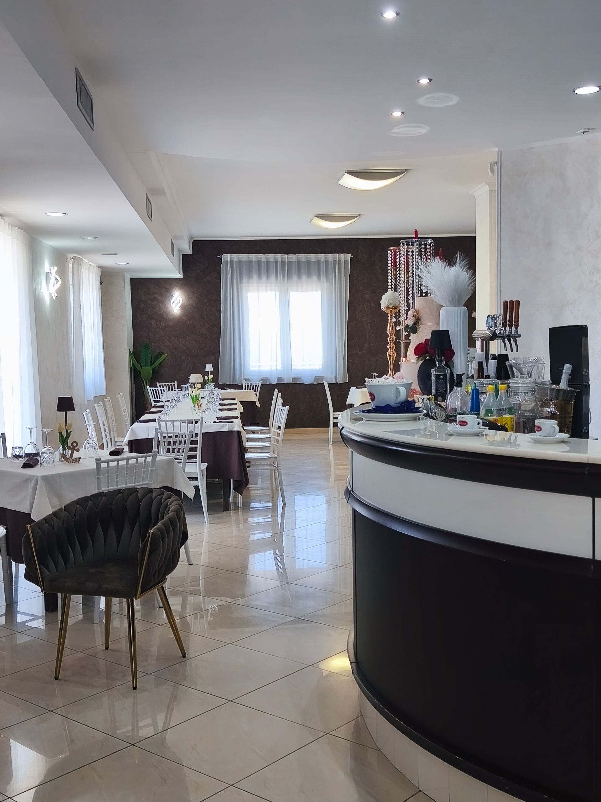 Luminoso interno del ristorante con tavoli apparecchiati, sedie nere e un bancone bar curvo bianco e nero.