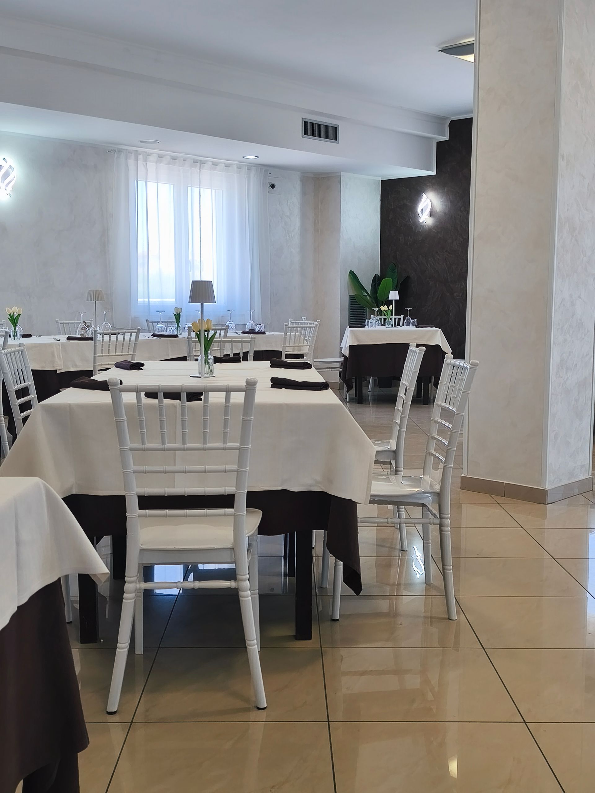 Elegante sala da pranzo con sedie bianche, tavoli apparecchiati e pavimento piastrellato.