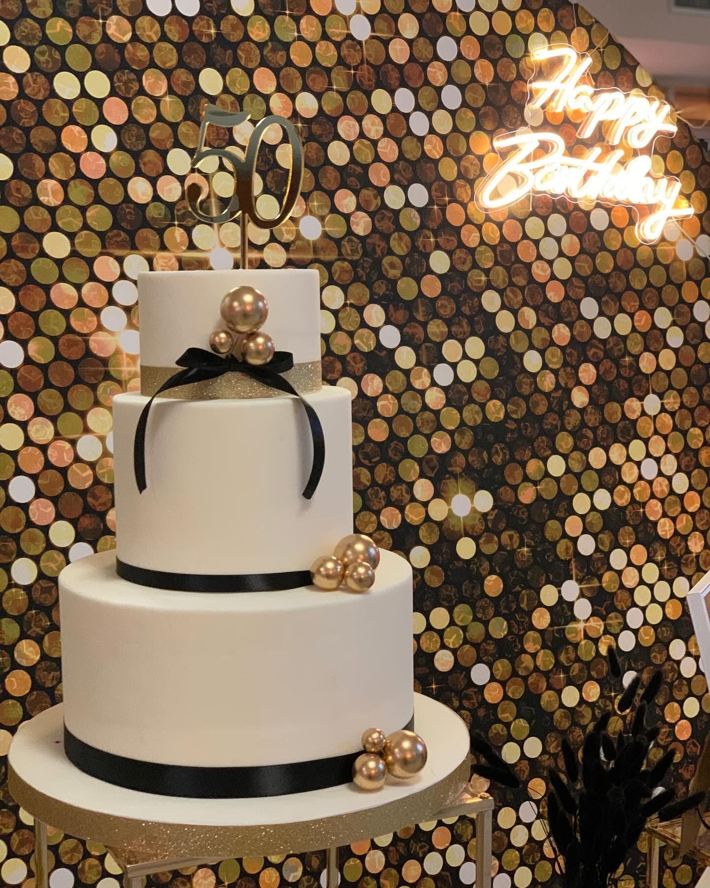 Torta a tre piani con decorazioni dorate, davanti a uno sfondo di paillettes dorate e un'insegna al neon per il compleanno.