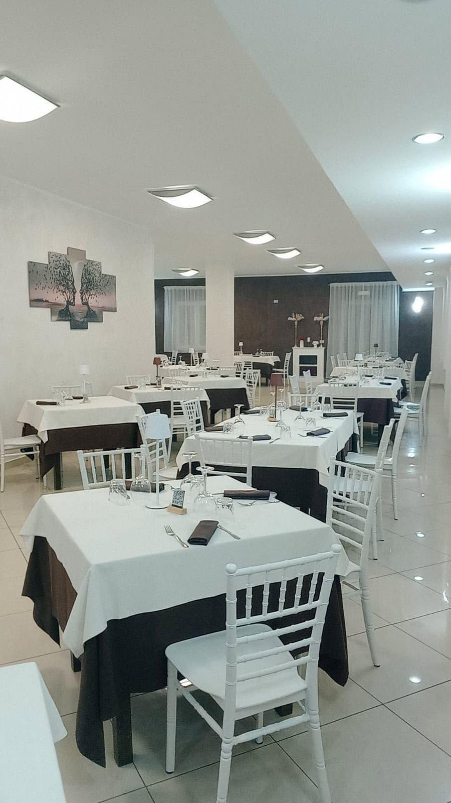 Sala da pranzo di un ristorante con tovaglie bianche, sedie bianche e luci intense sul soffitto.
