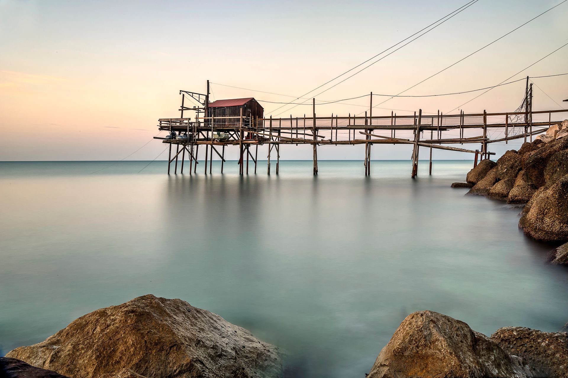 Un pontile di legno per la pesca su acque calme al tramonto, con rocce in primo piano e un cielo dai toni pastello.