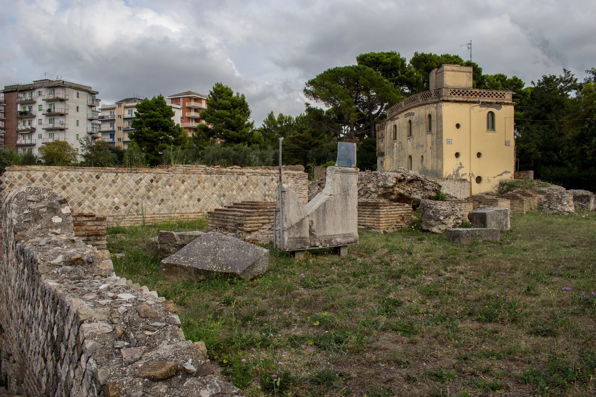 Rovine in pietra e muretti bassi in un campo erboso, con palazzi e cielo nuvoloso sullo sfondo.