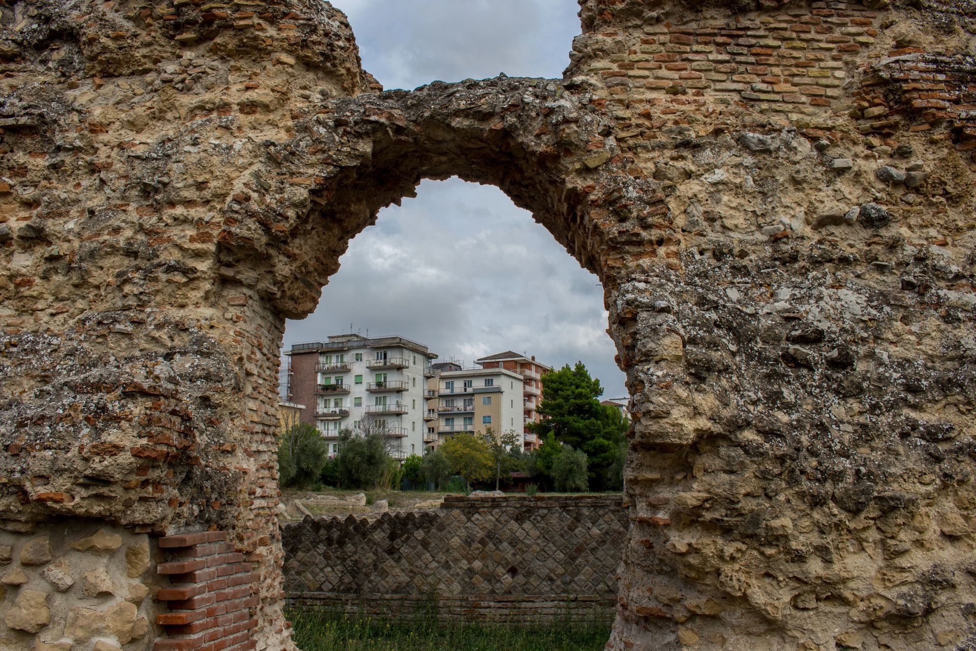 Rovine di un muro di pietra che incorniciano palazzi residenziali sotto un cielo nuvoloso.