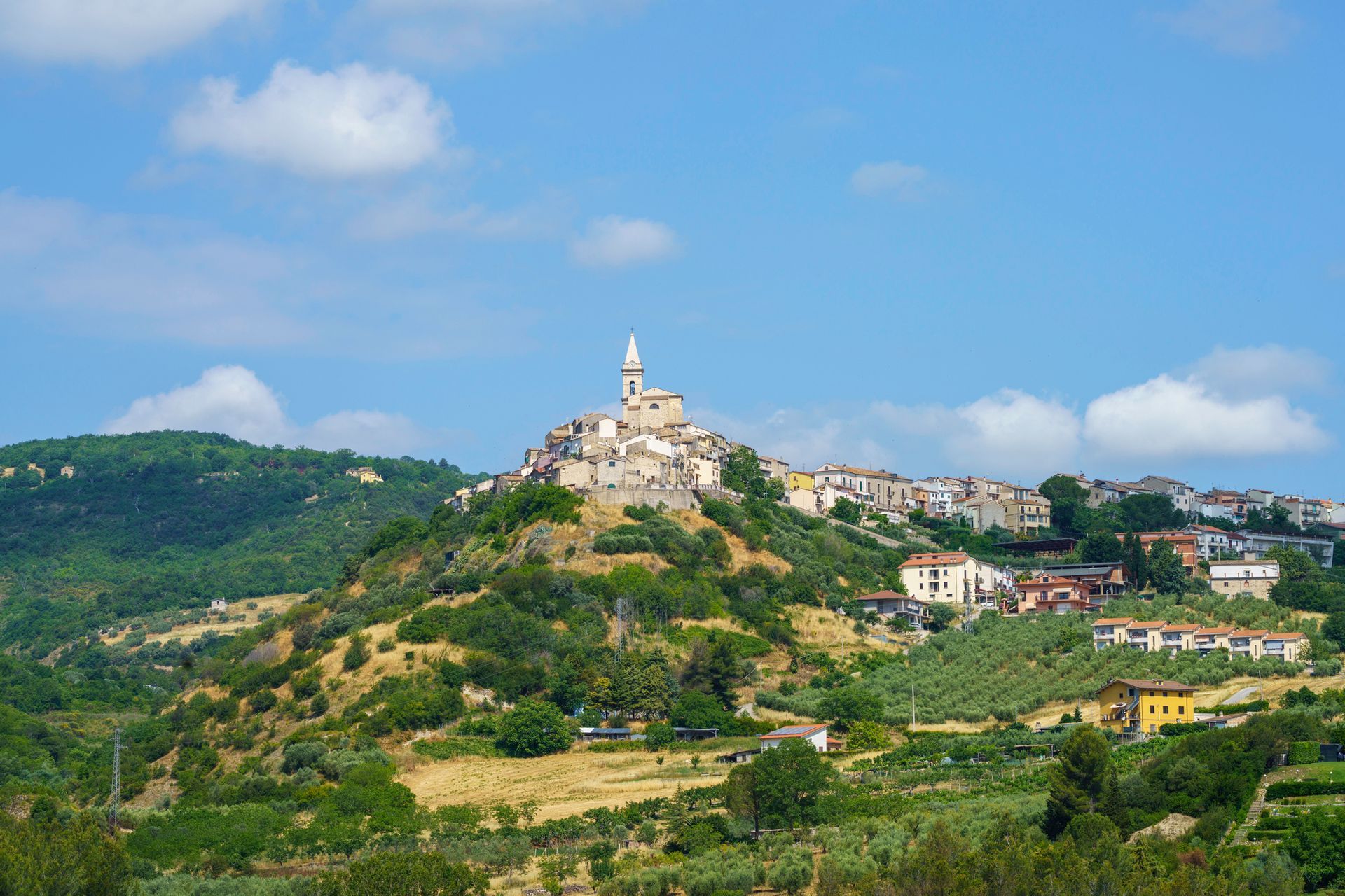 Villaggio collinare con il campanile bianco che svetta su verdi colline sotto un cielo azzurro.