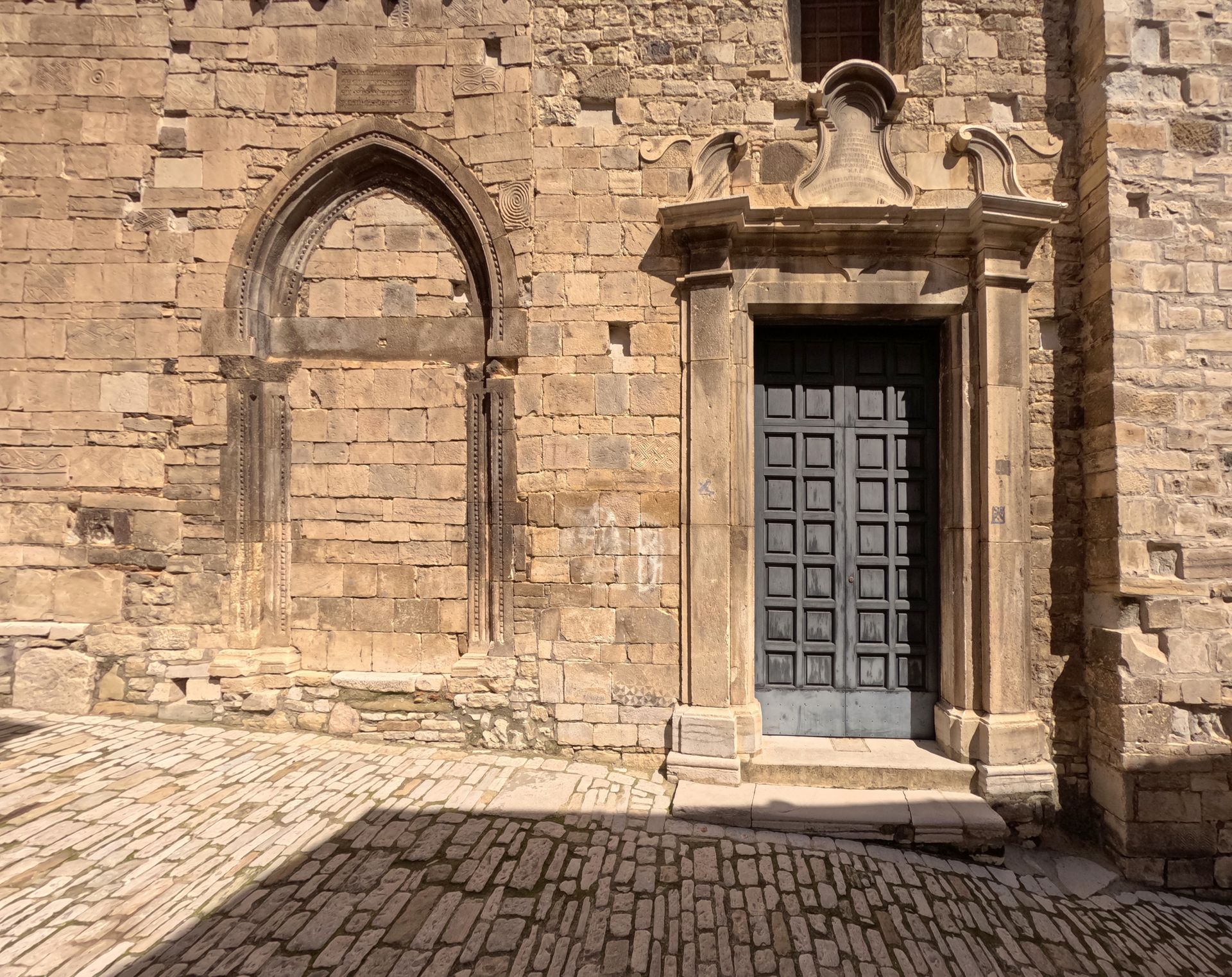 Facciata di un edificio in pietra con finestra ad arco, portale decorato e pavimentazione in ciottoli illuminata dal sole.