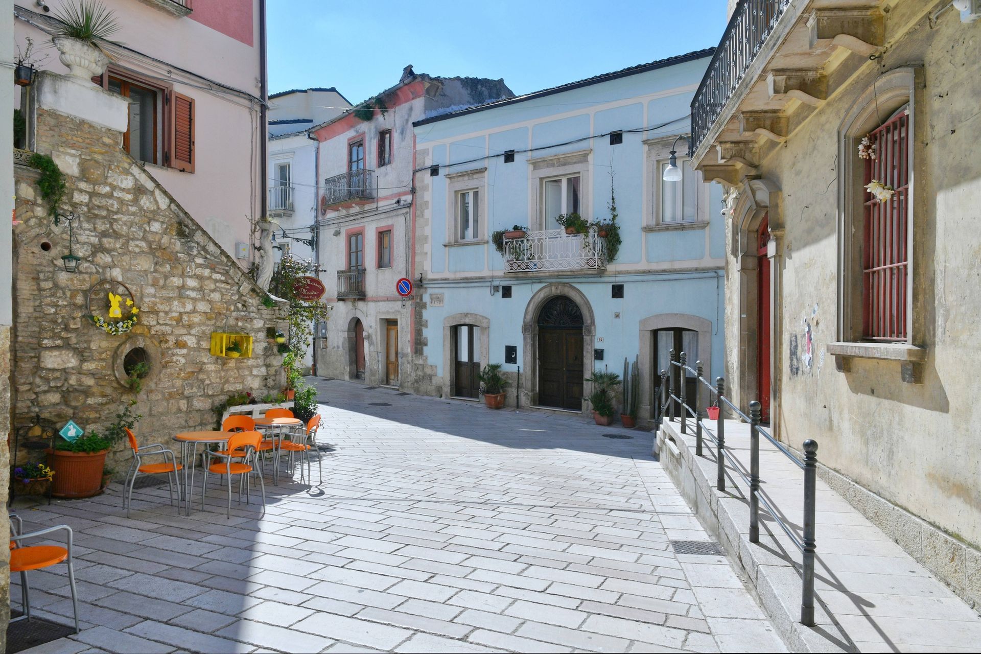 Una strada acciottolata e soleggiata, con edifici bianchi, muri in pietra e sedie arancioni da caffè, in una tranquilla piazza di paese.
