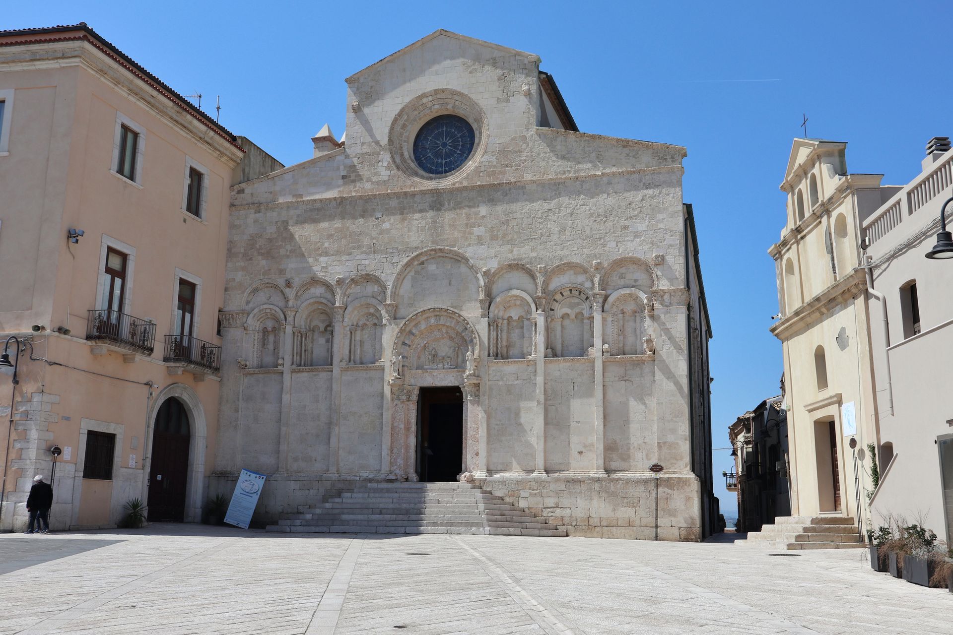 Facciata in pietra della cattedrale su una soleggiata piazza lastricata, fiancheggiata da edifici beige sotto un cielo azzurro.