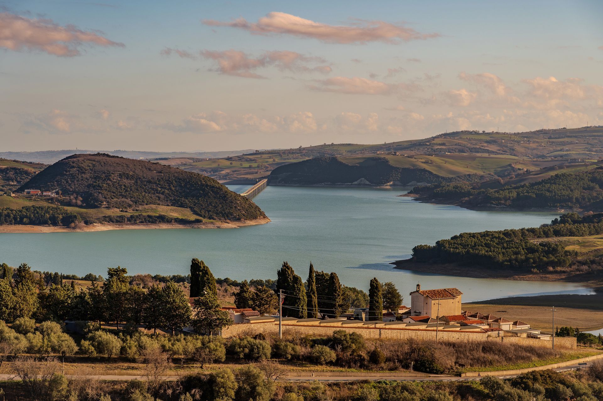 Ampio lago incastonato tra dolci colline sotto un cielo parzialmente nuvoloso, con alberi e campi in primo piano.