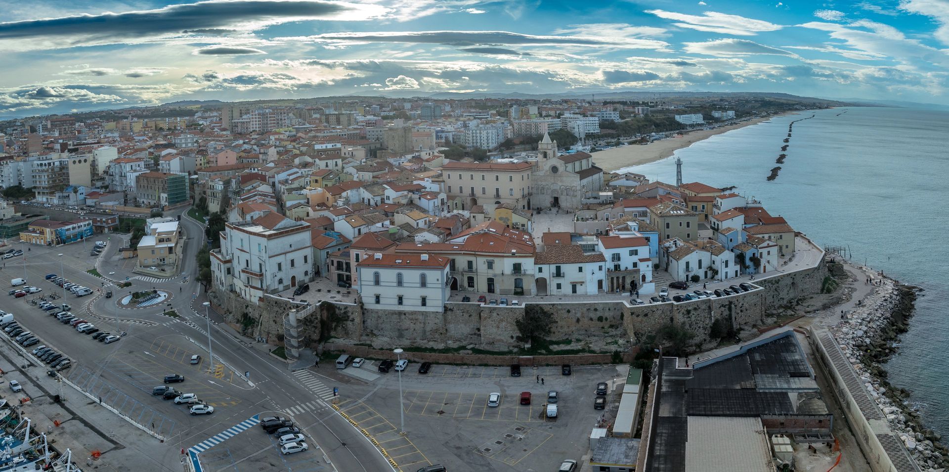 Veduta aerea di una cittadina costiera con edifici dai tetti rossi, un porto e una costa rocciosa sotto un cielo nuvoloso.