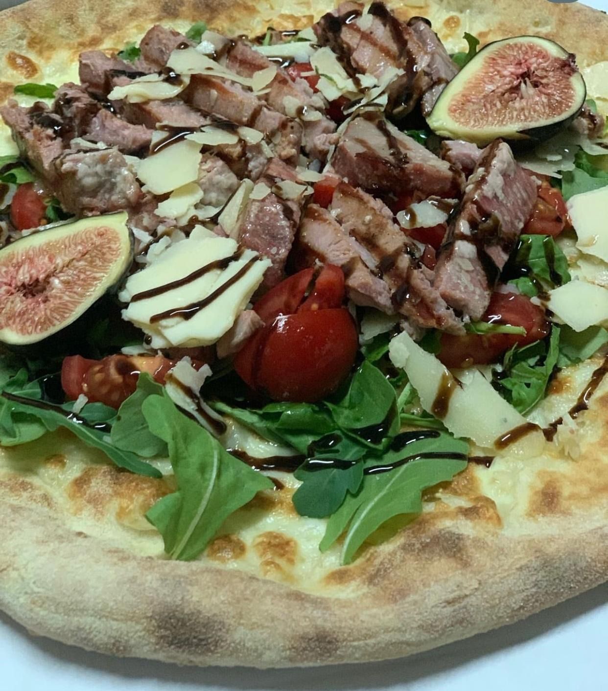 Pizza su pane piatto condita con rucola, carne a fette, fichi, formaggio grattugiato, pomodorini e glassa balsamica.