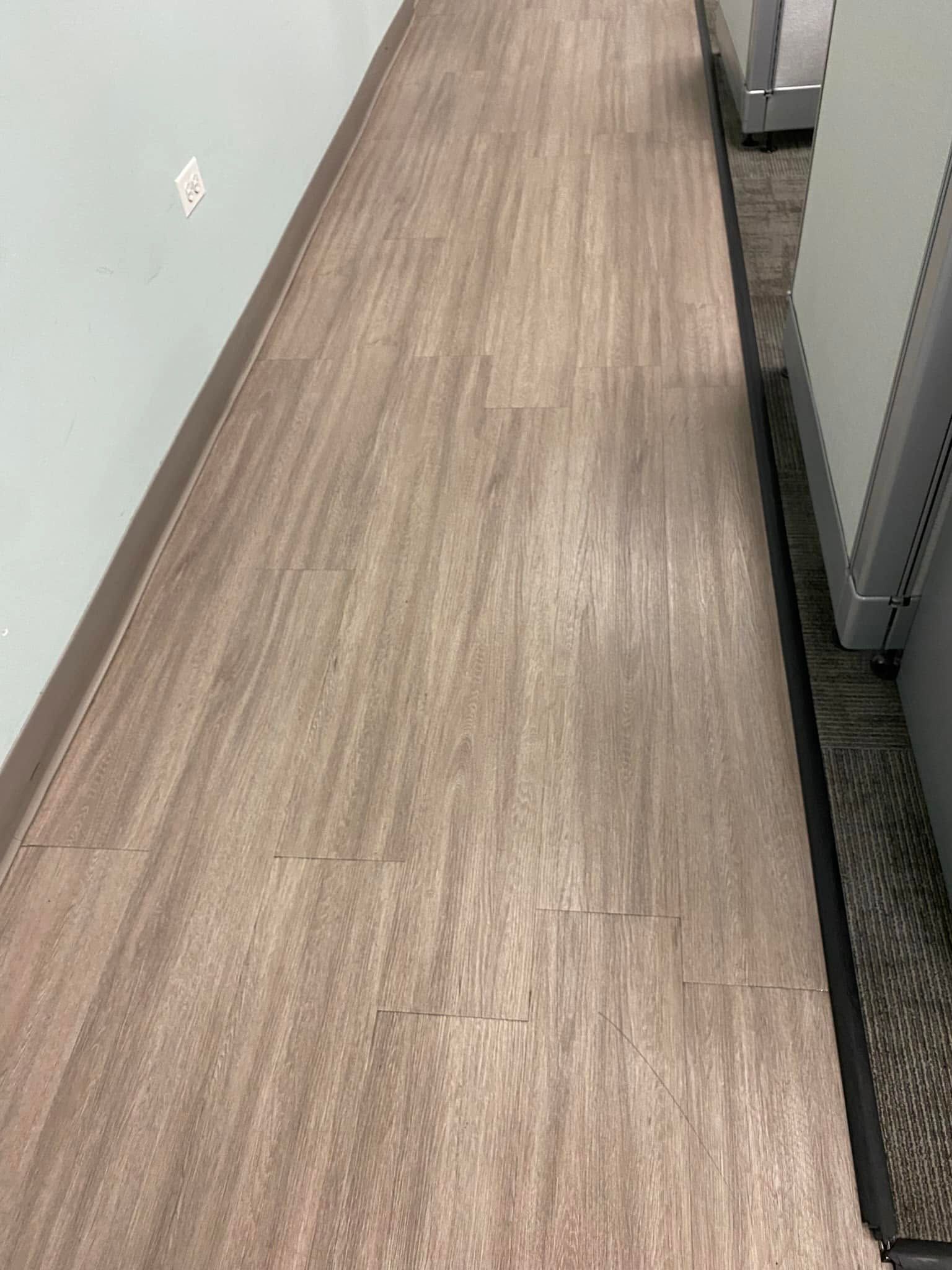 Narrow office hallway with wood-look flooring and light gray walls, viewed from above.