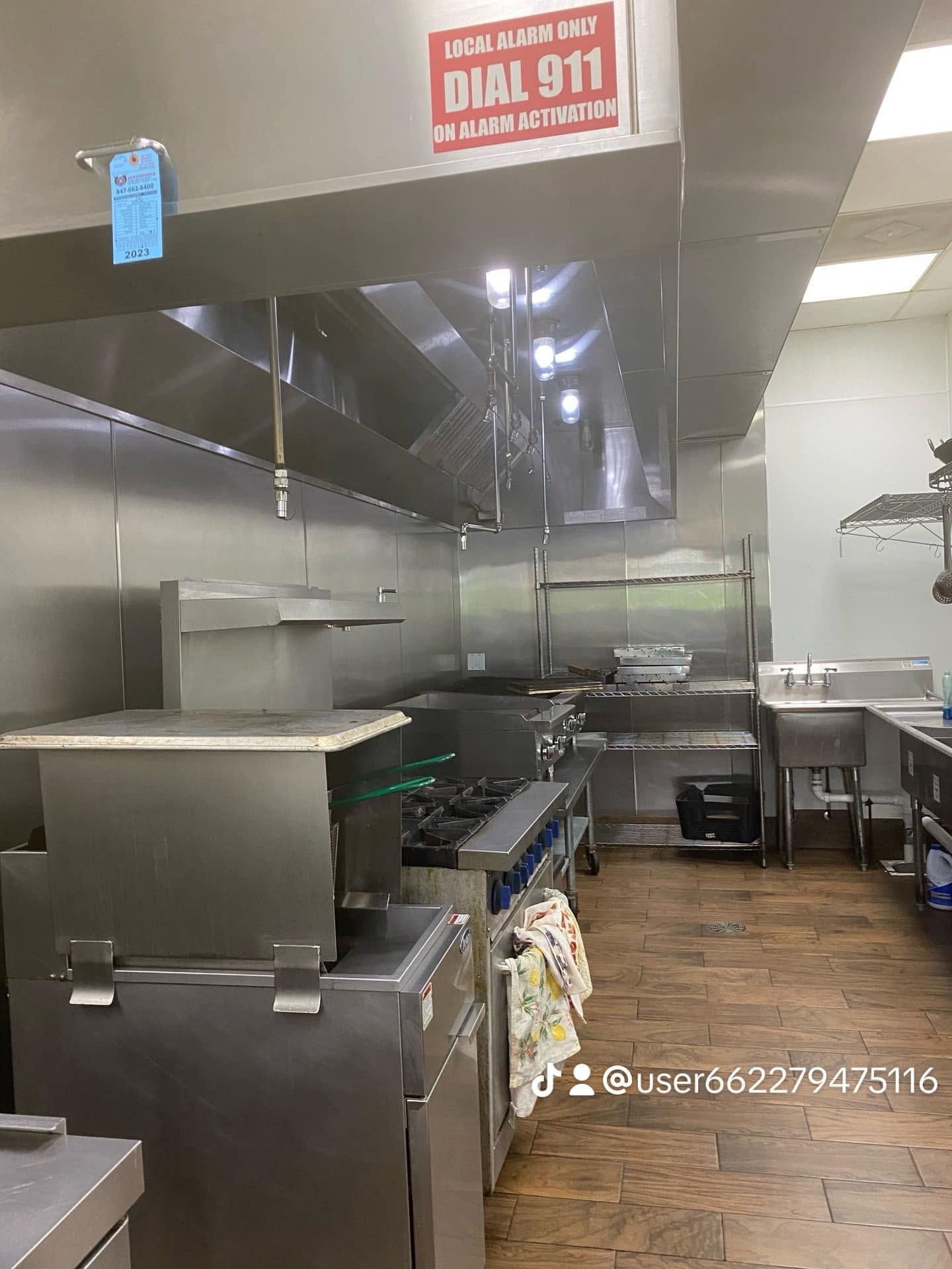 Commercial kitchen with stainless steel equipment, prep counters, and a sink under bright overhead lights.