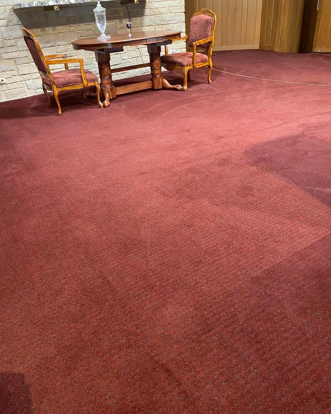 Empty room with burgundy carpet, two chairs, and a small table near a stone fireplace.