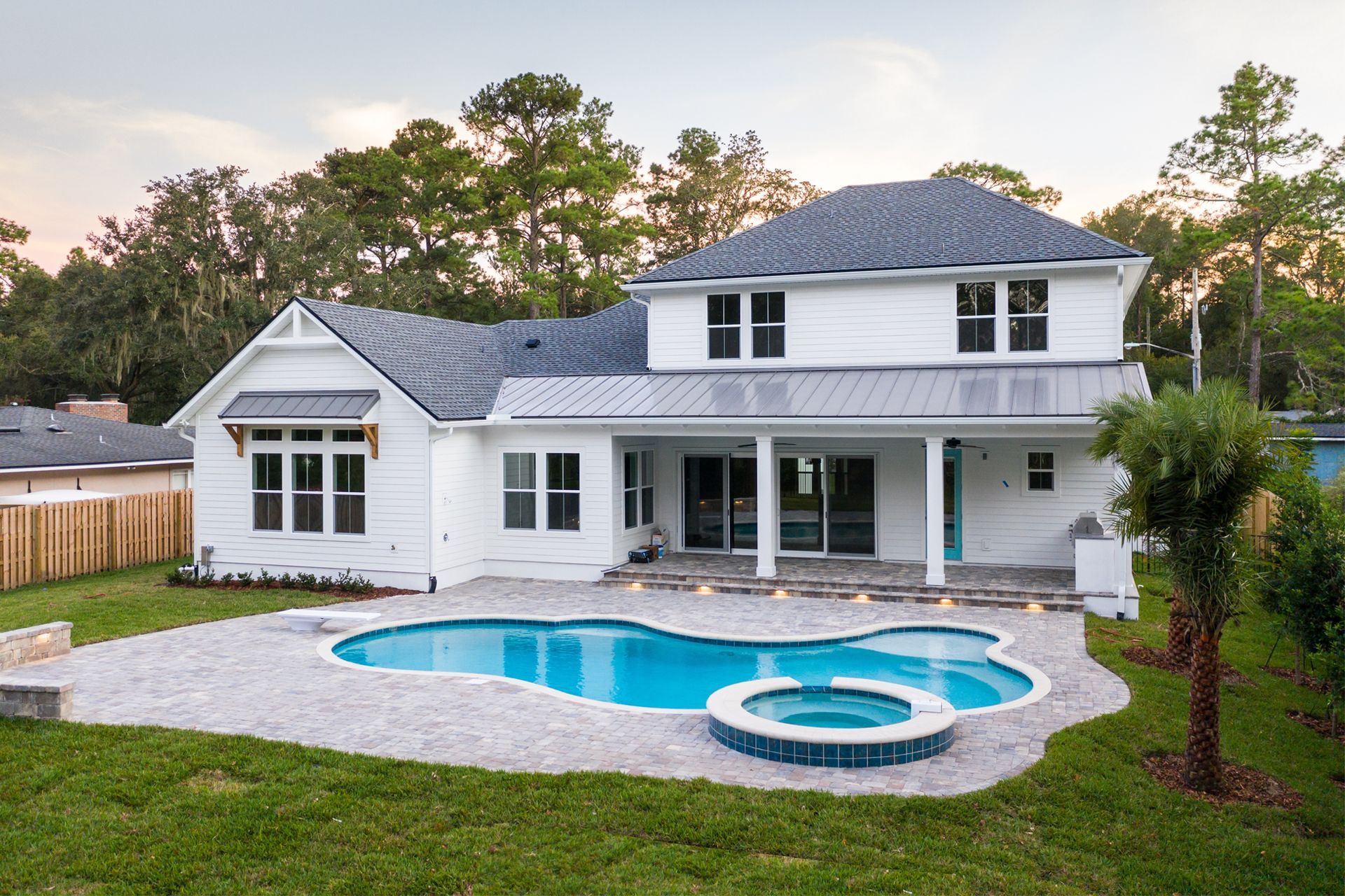 White house with blue pool and spa, patio, and lawn. Trees in the background.