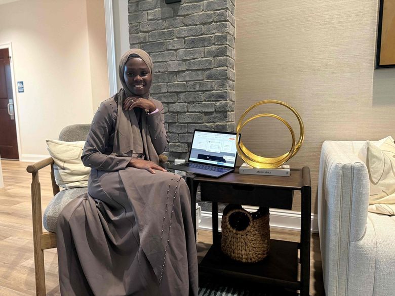 Woman seated beside a laptop on a small table in a lounge, with decorative rings and a basket nearby.