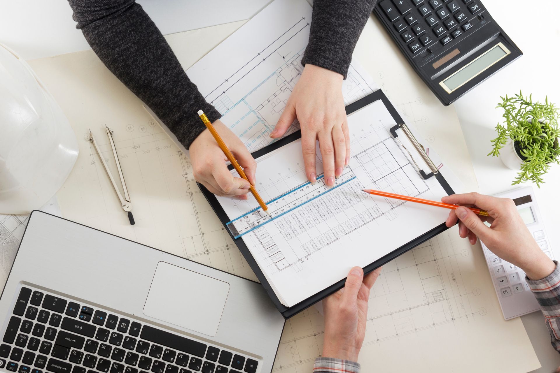 Hands planning on a desk with a clipboard, laptop, ruler, and calculator around charts and graphs.