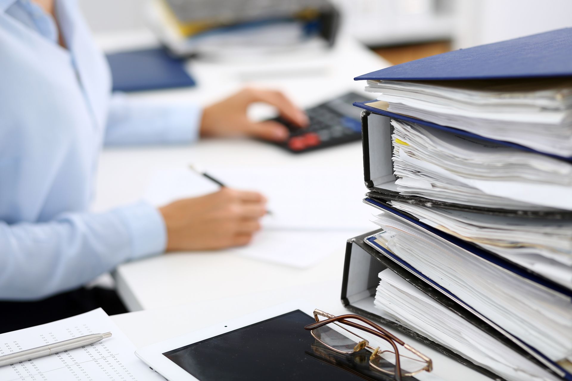 Office desk with stacked binders and documents beside a person writing on paperwork