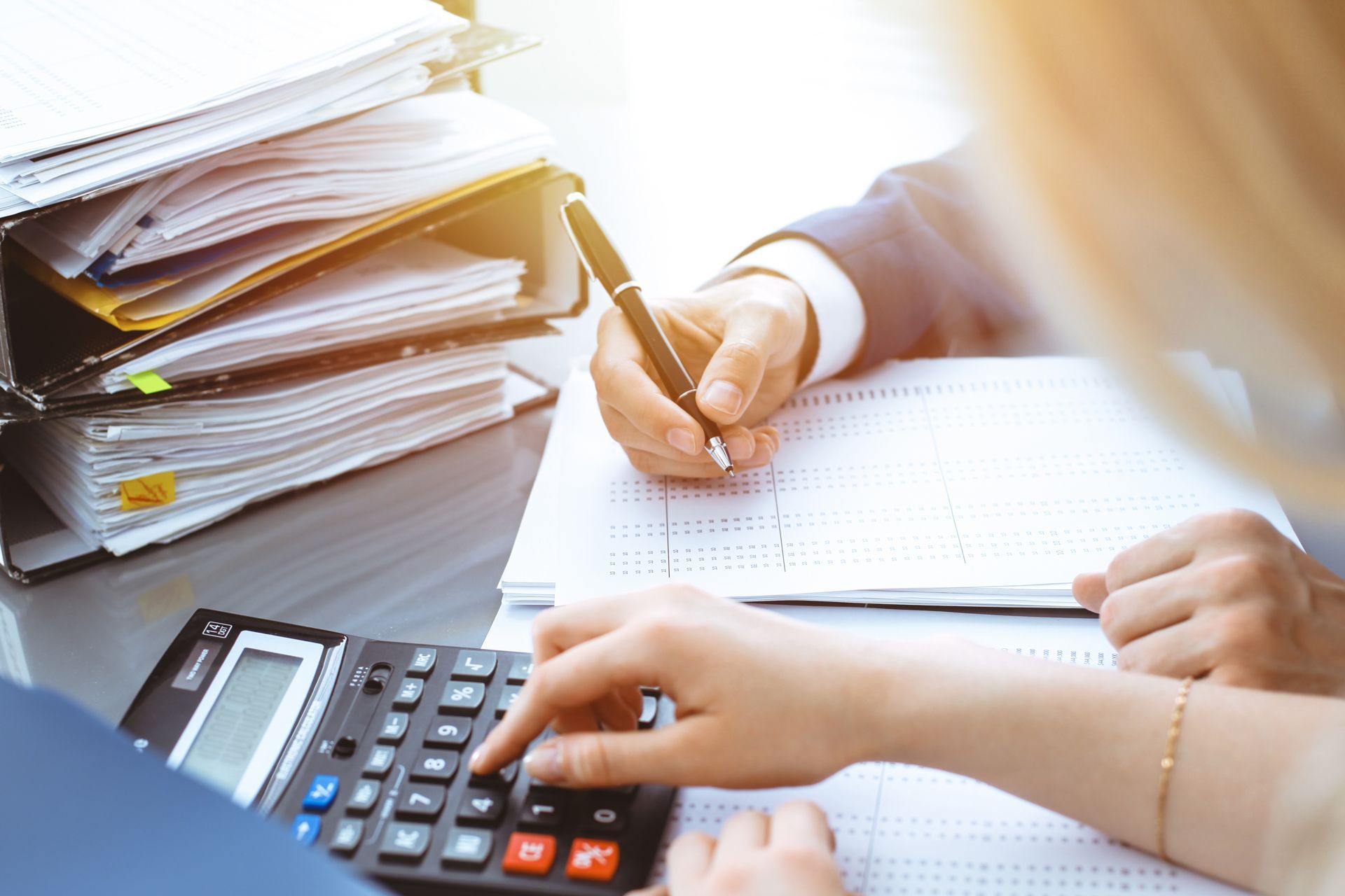 Hands writing in a notebook beside a calculator and stacked papers in a bright office setting