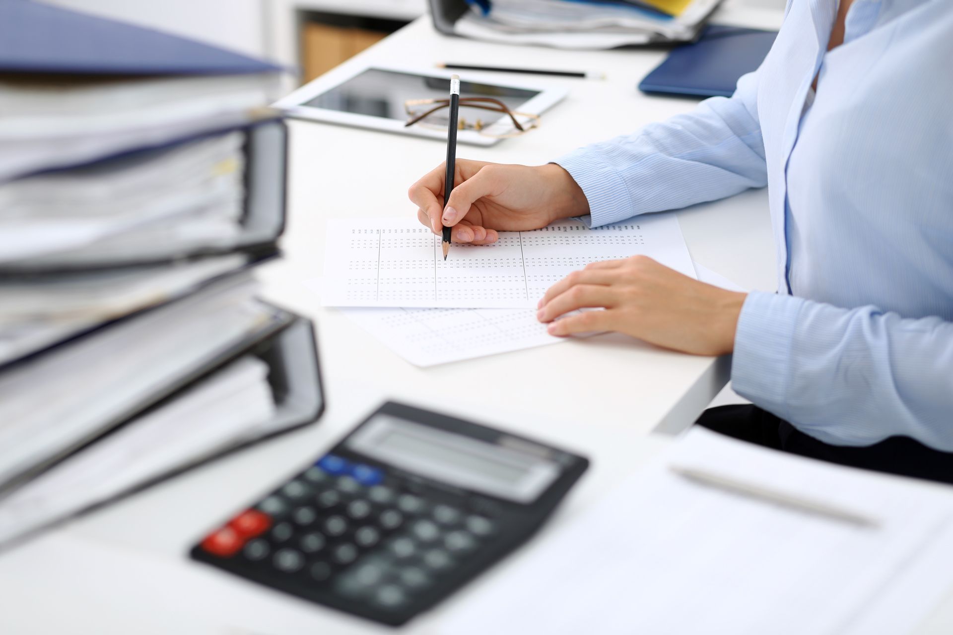 Hands writing on papers at an office desk with a calculator and stacked files