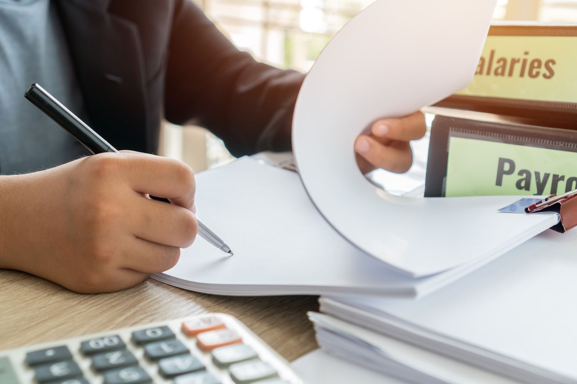 Hands using calculator and pen over financial documents, with payroll folders in the background