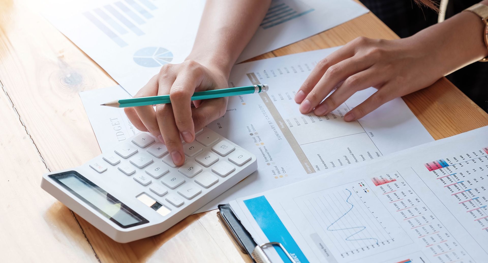 Hands using a calculator and pencil over financial charts and papers on a desk