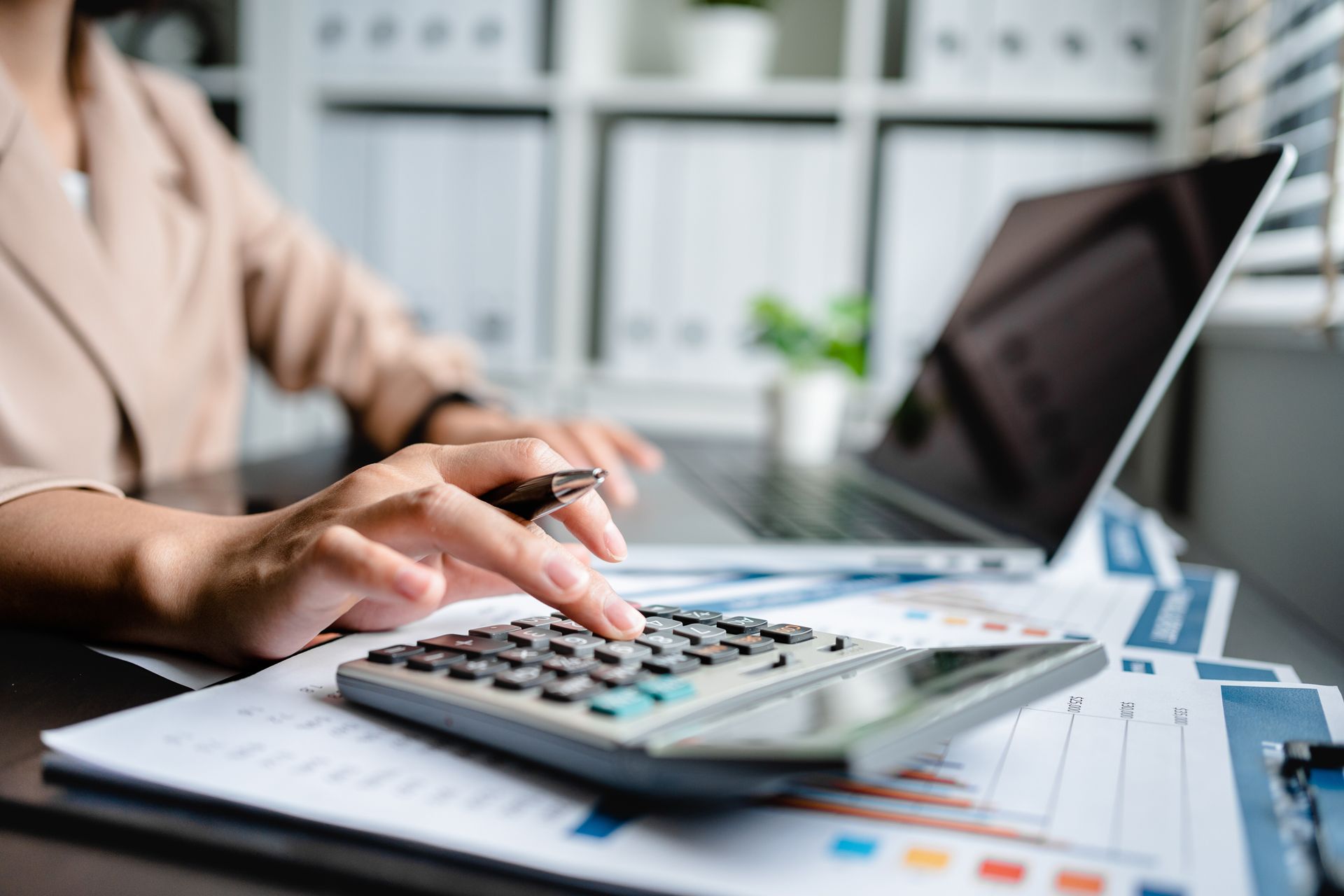 Hands using a calculator beside a laptop and paperwork on a desk