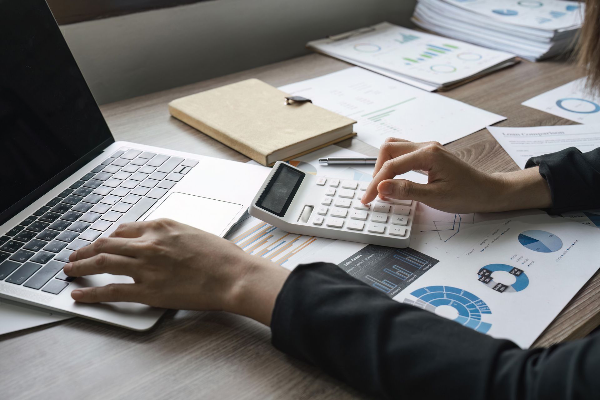 Hands using a laptop and calculator on a desk with paperwork and charts
