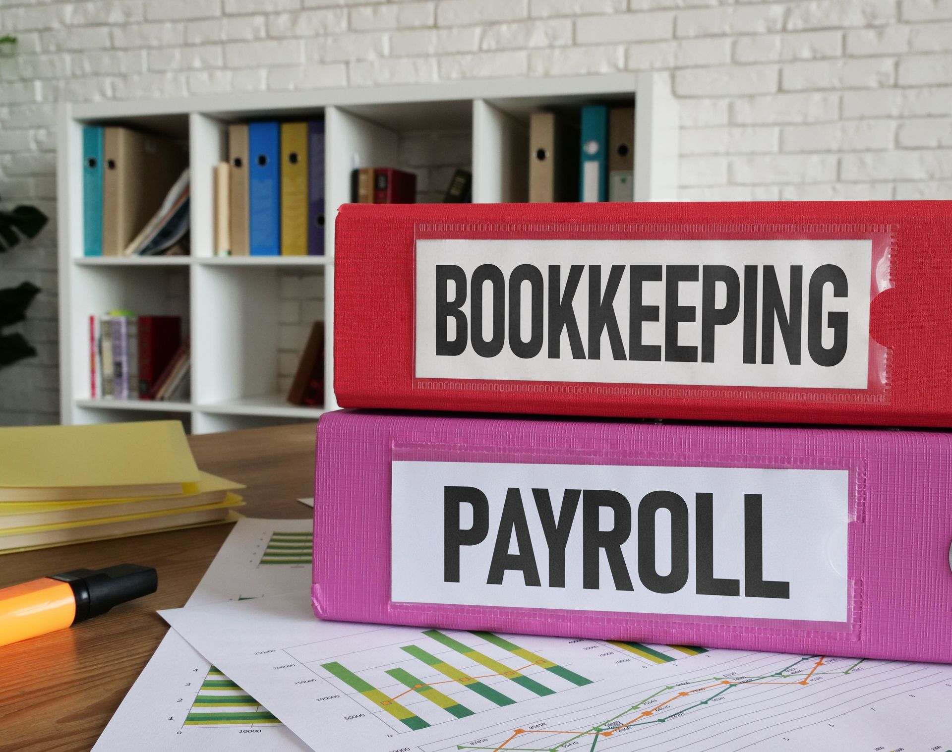 Pink and red binders labeled “BOOKKEEPING” and “PAYROLL” on a desk with charts and office supplies.