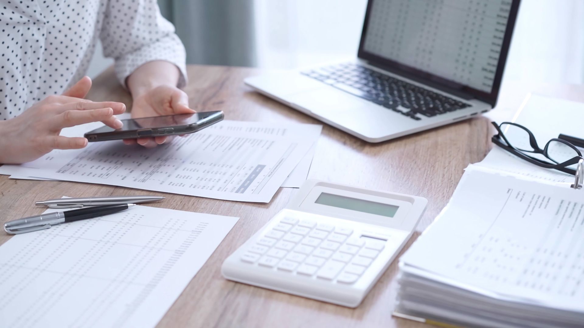Person using a smartphone at a desk with paperwork, calculator, laptop, and glasses.