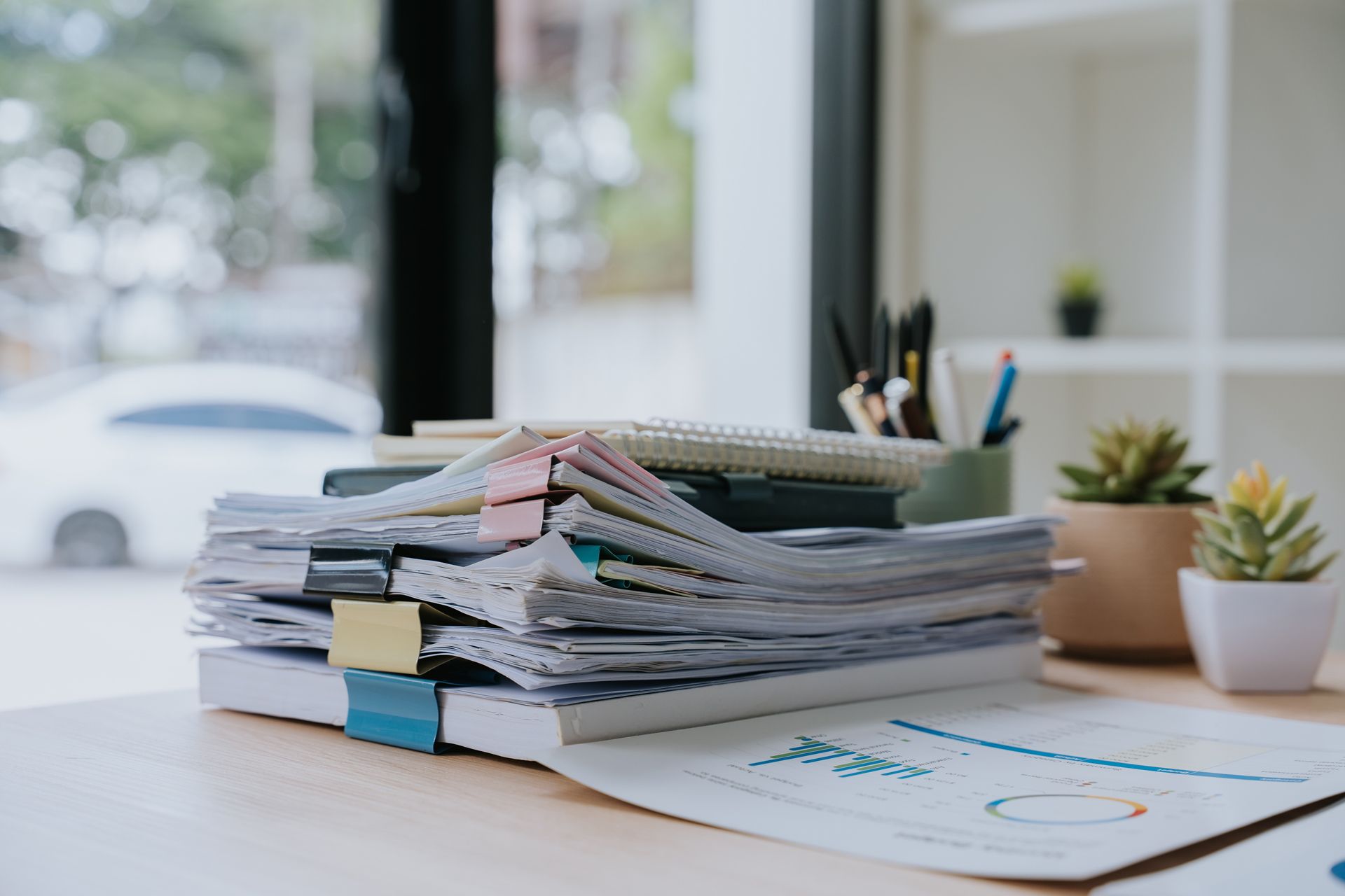 Stack of paper documents with clips on a desk beside a notebook and pen cup by a window