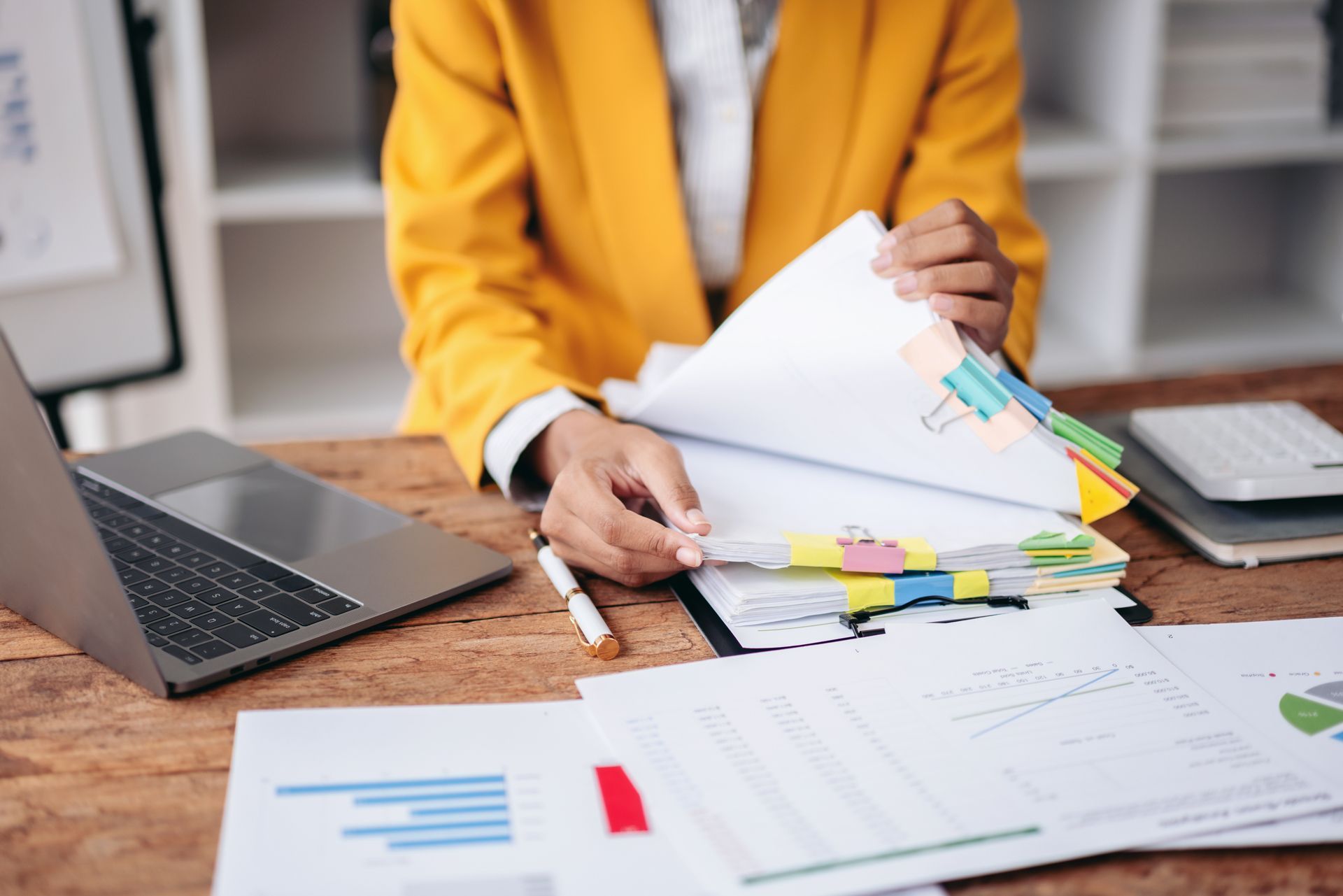 Person in a yellow blazer reviewing documents at a desk with a laptop and sticky notes.