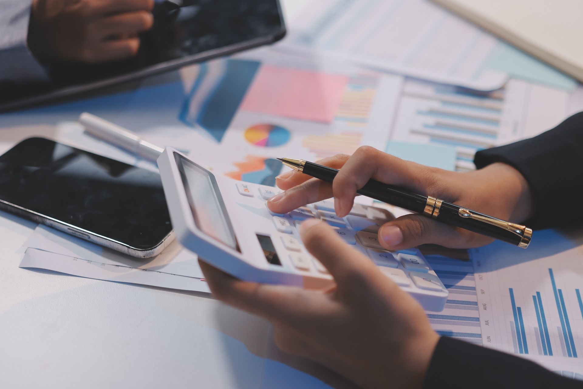 Hands reviewing business charts on a tablet with a pen on a desk, with a notebook and phone nearby.
