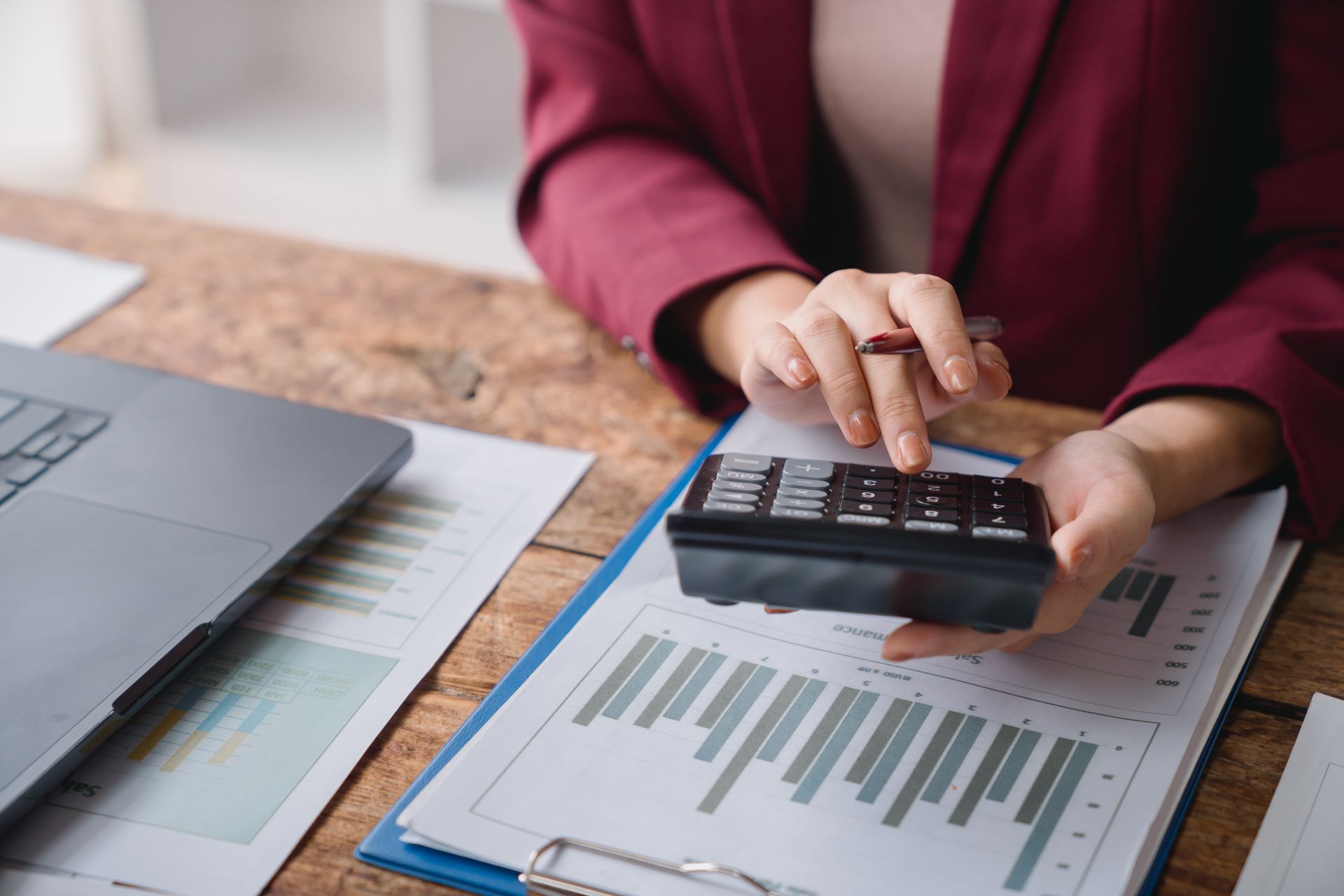 Hands using a calculator over financial charts beside a laptop on a desk