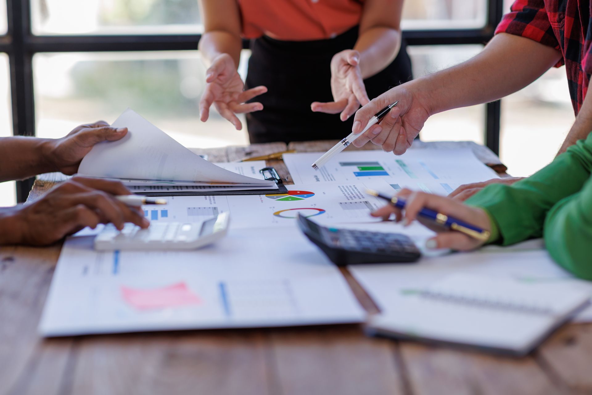 People gathered around a table reviewing papers, charts, and a calculator in a collaborative meeting.