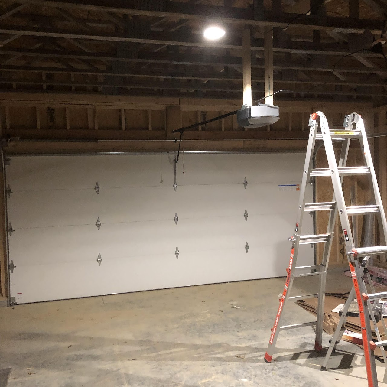 White garage door inside a garage under construction, with a ladder and garage door opener.