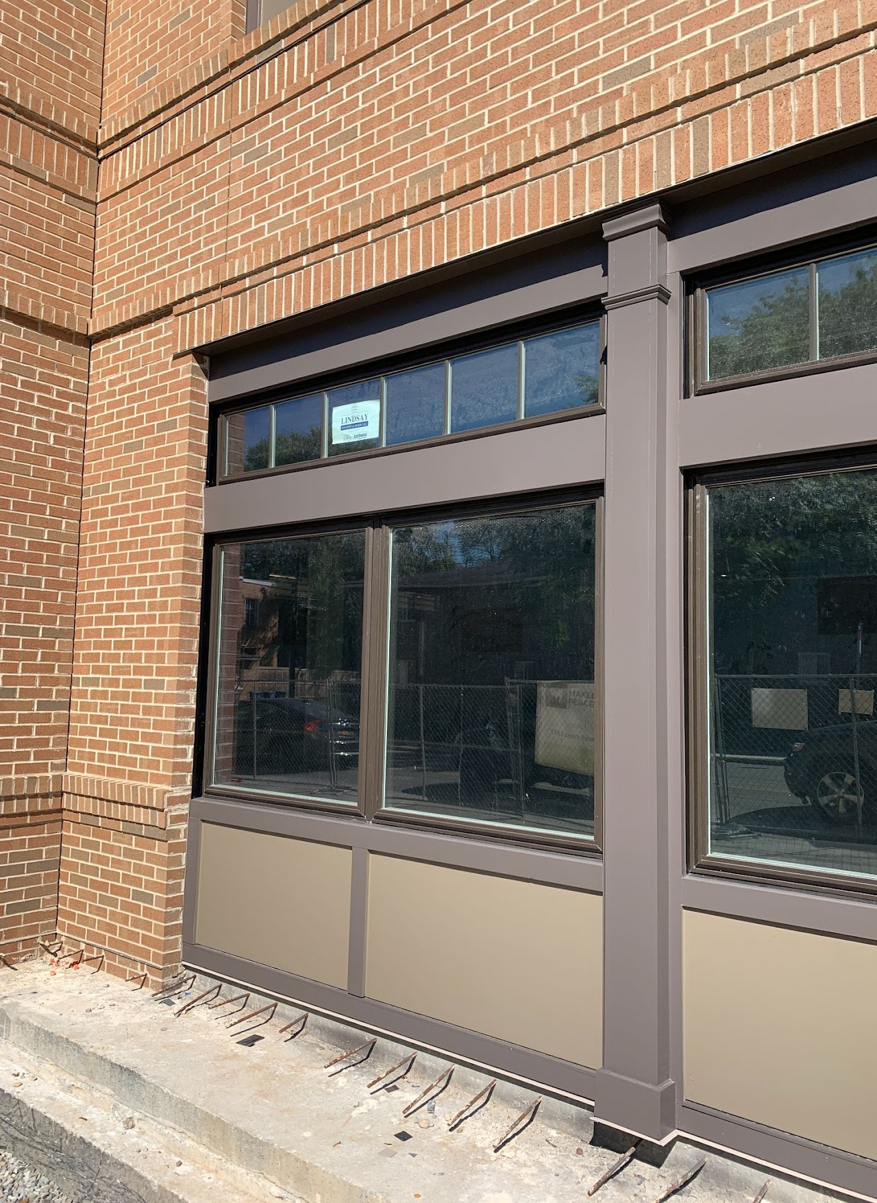 Exterior view of a brick building with large brown-framed windows, tan panels below, and a sidewalk in front.