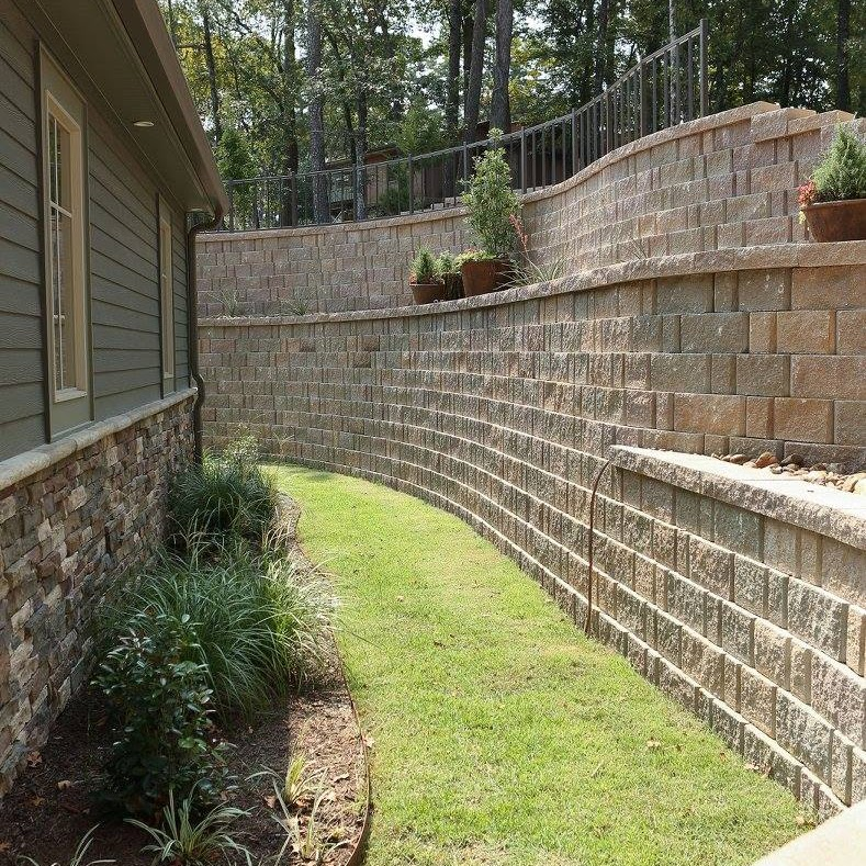 Curved retaining walls made of stone blocks alongside a narrow grassy area, next to a house.