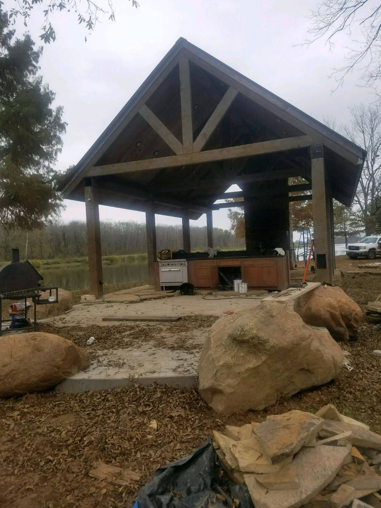 A wooden pavilion with a kitchen area, near a lake. Large rocks sit in the foreground.