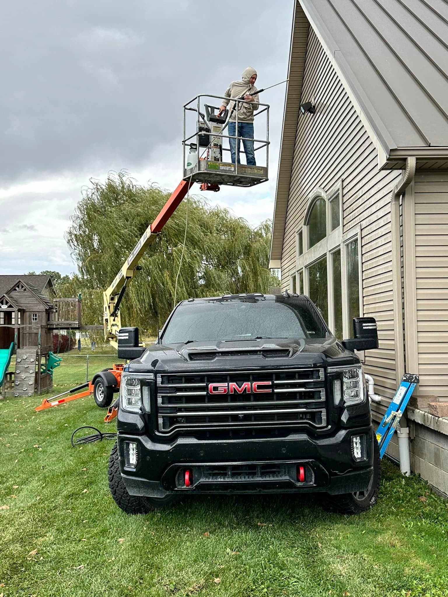 A man in a lift basket works on the side of a house. A black GMC truck is parked on the lawn.