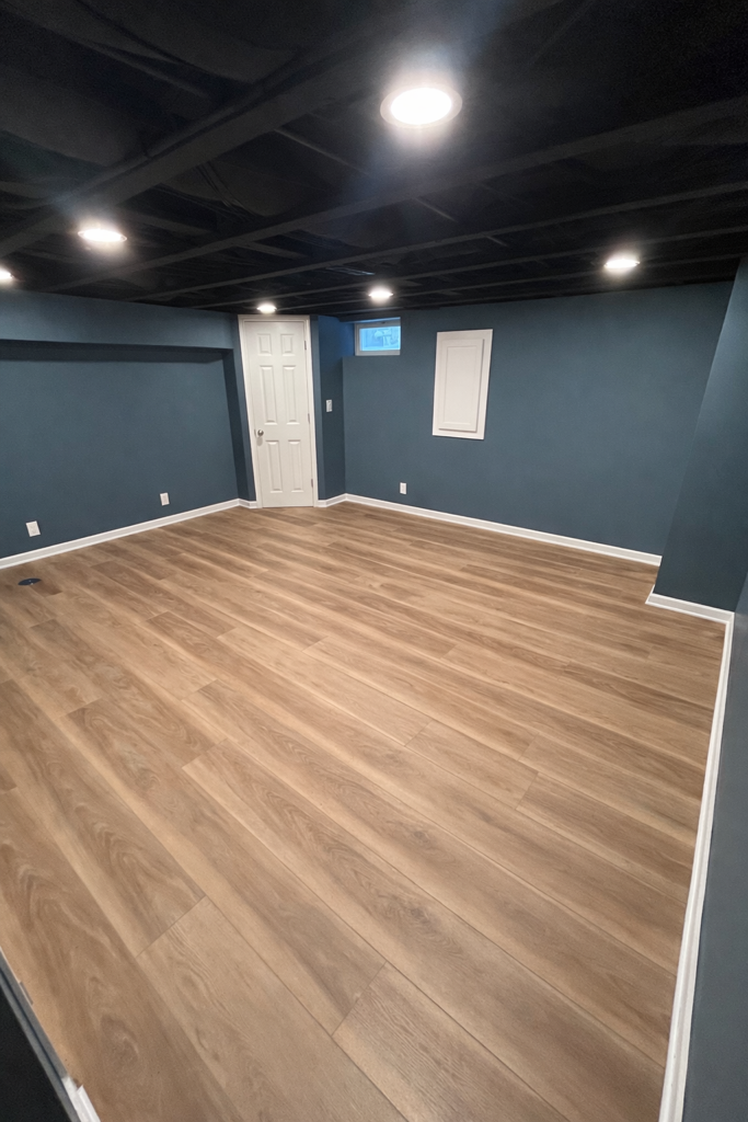 An empty basement room with dark blue walls, a black painted ceiling, recessed lighting, and wood-look laminate flooring.