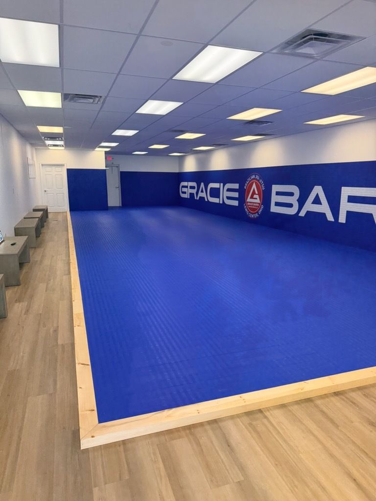 Empty Brazilian jiu-jitsu mat room with blue mats, white ceiling lights, and wall logo reading “GRACIE BARRA.”