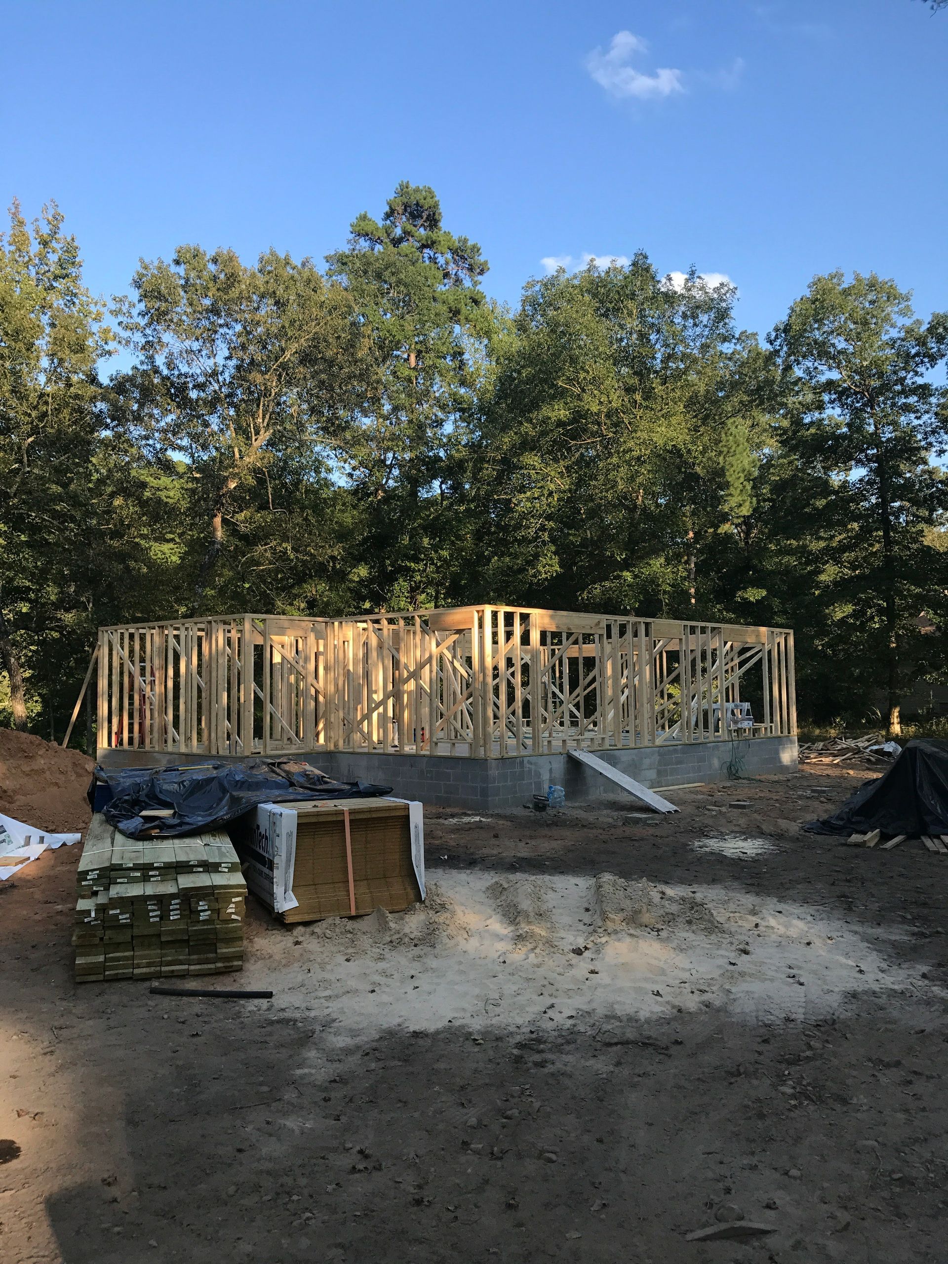 Framed wooden structure of a house under construction. Lumber and foundation visible with trees in the background.