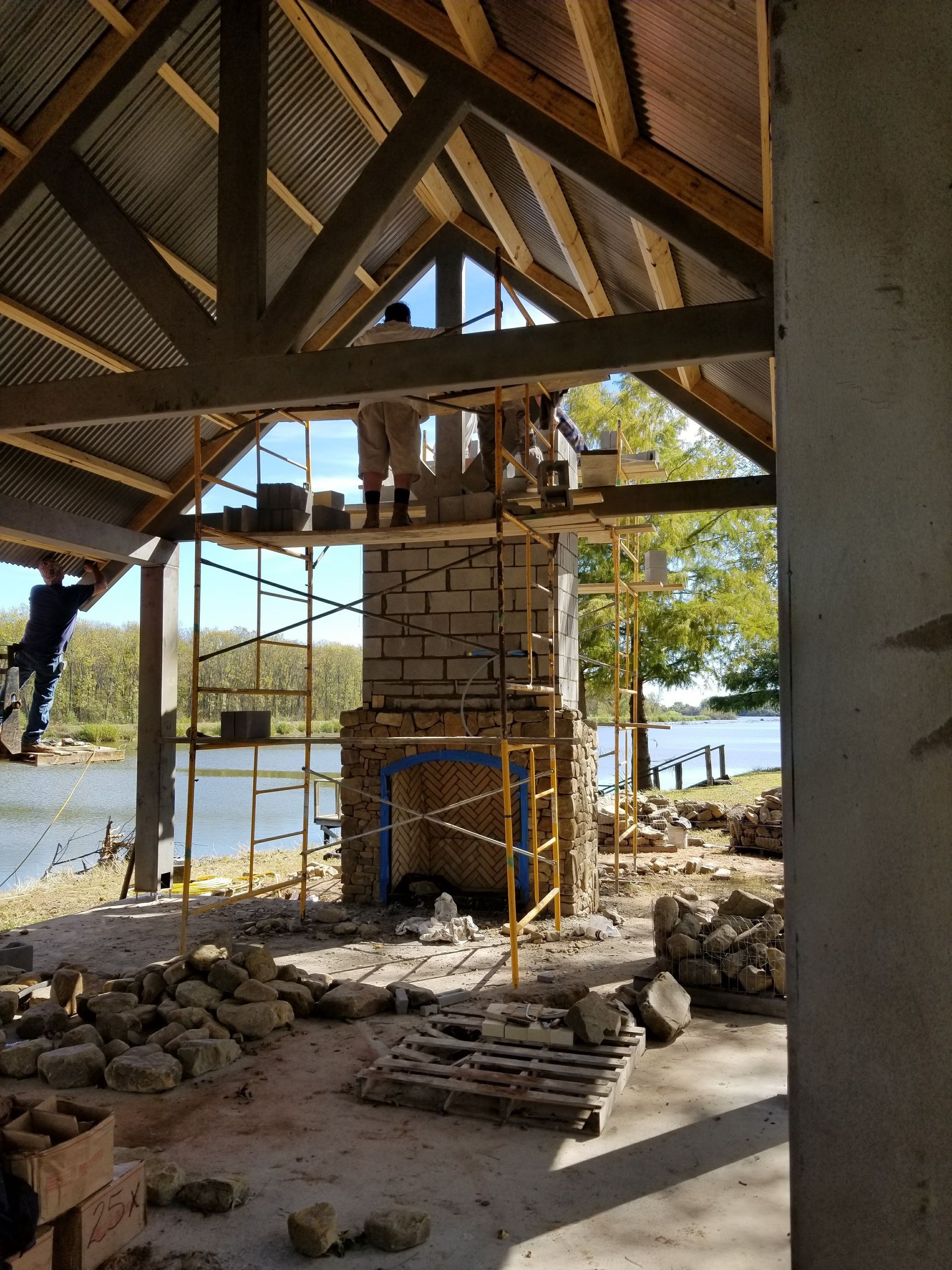 Construction of a stone chimney in a waterfront pavilion; workers on scaffolding, blue sky and water in the background.