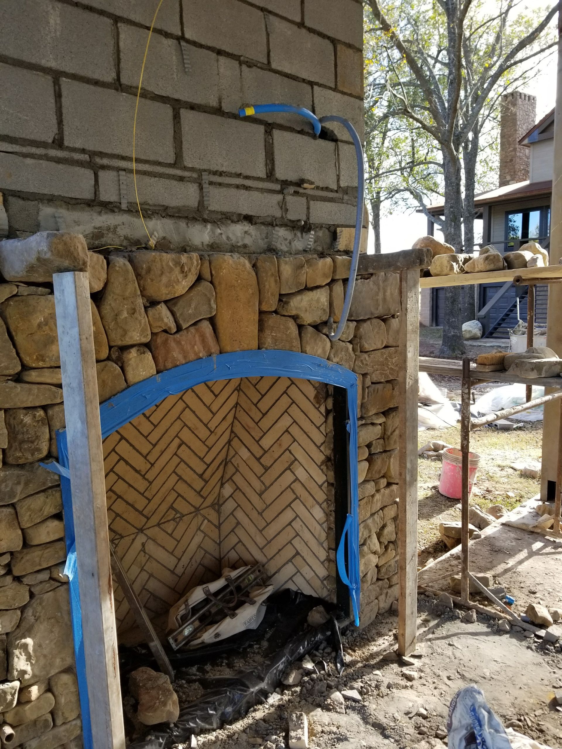 Outdoor fireplace under construction with stone and brick, blue trim, and utility lines.