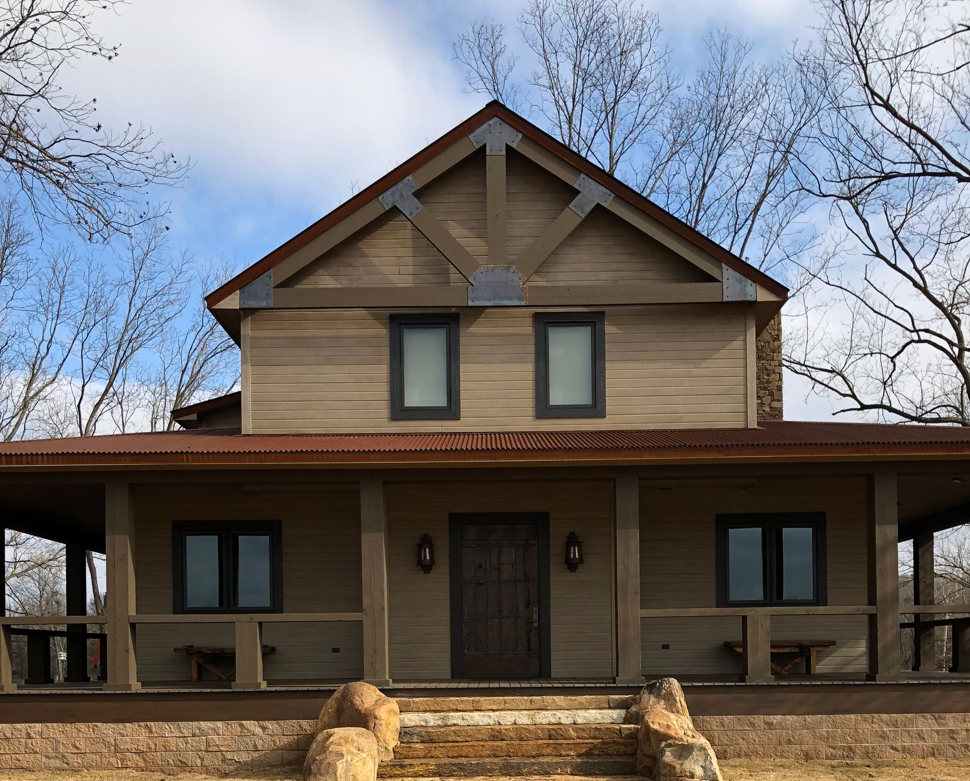 Rustic two-story house with brown siding, porch, and a metal roof. Blue sky with bare trees in the background.