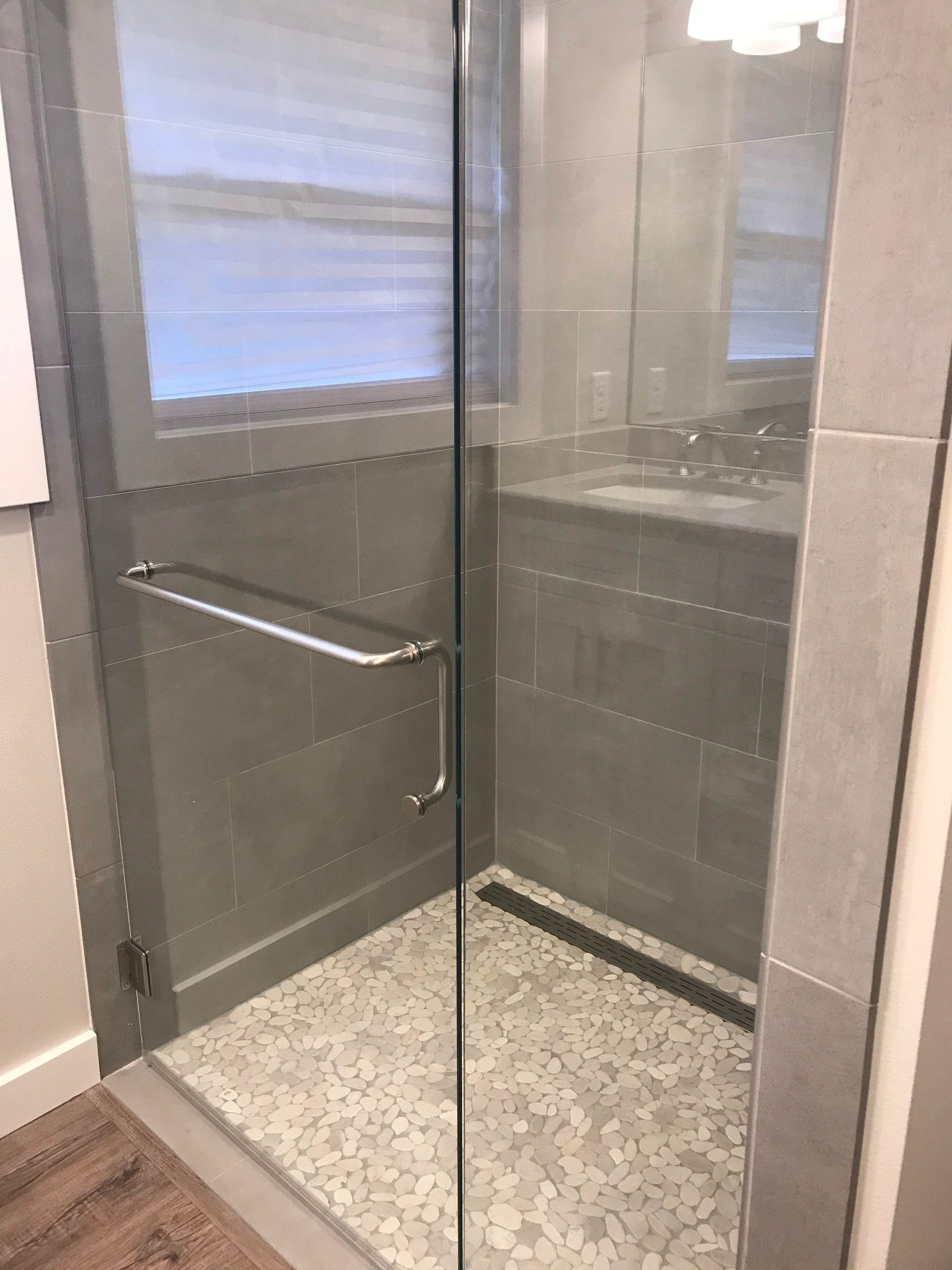Glass shower enclosure with gray tiled walls and pebble floor. Chrome door handle.